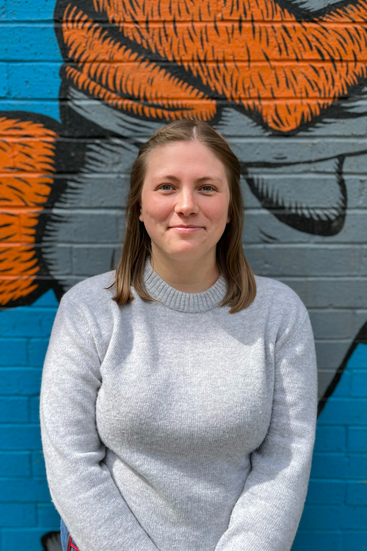 a photo of Alice, our Director. She is stood in front of an orange and blue painted wall. She is smiling at the camera