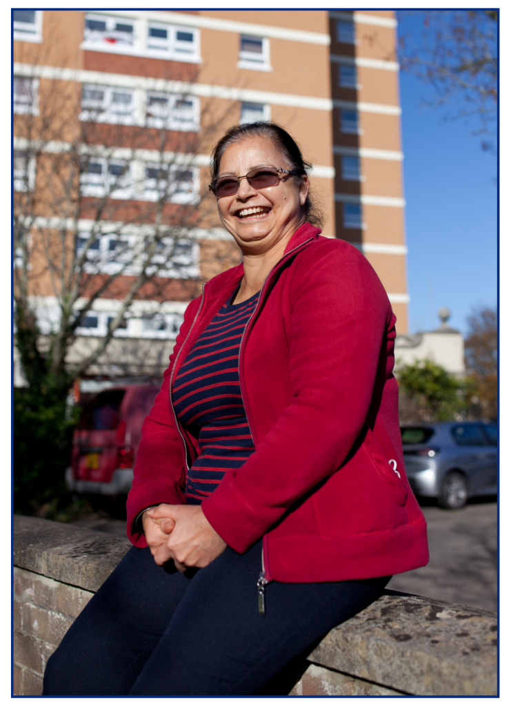 A woman sat on a wall outside a tower block of flats. She is laughing.