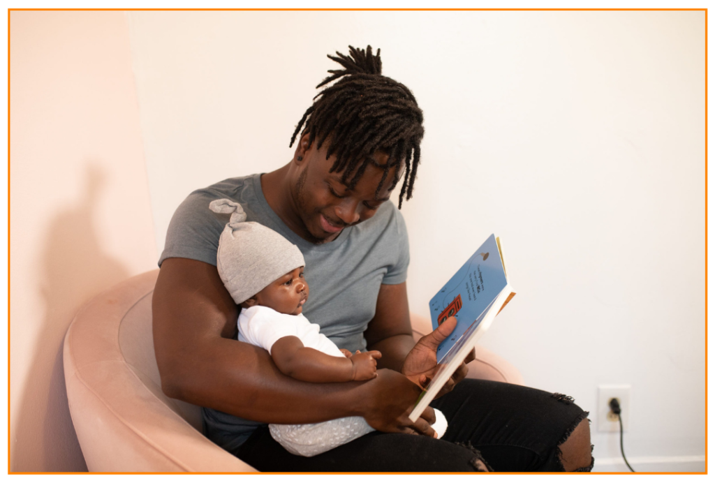 A man sat in an armchair reading a book with his baby who is sat on his lap.