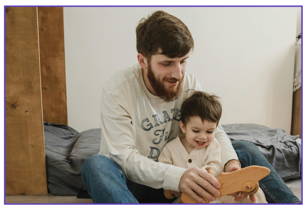 Photograph of a man sat on the edge of a bed playing with a toy with his toddler.