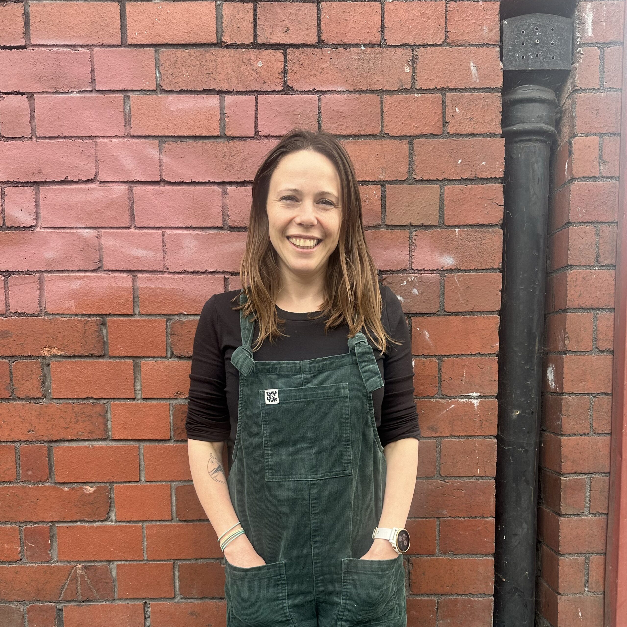A photo of Daisy stood in front of a red brick wall. She is smiling at the camera.